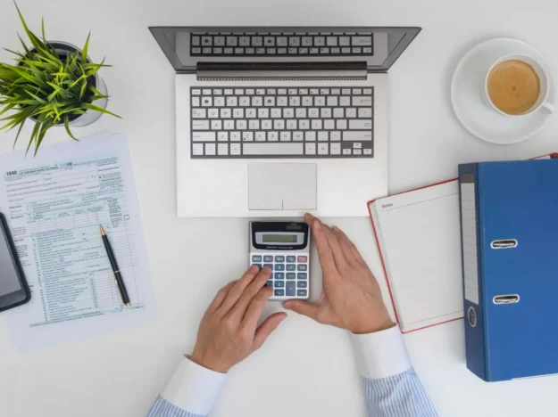 Vue de dessus d'un bureau avec un ordinateur, une calculatrice, des documents fiscaux et un classeur, symbolisant la gestion et le calcul des impôts professionnels.