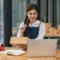 Jeune entrepreneure e-commerce souriante tenant un colis et travaillant sur son ordinateur portable, entourée de cartons prêts à être expédiés. Illustration de la gestion des commandes et de l'importance des conditions générales de vente (CGV) pour sécuriser les transactions.