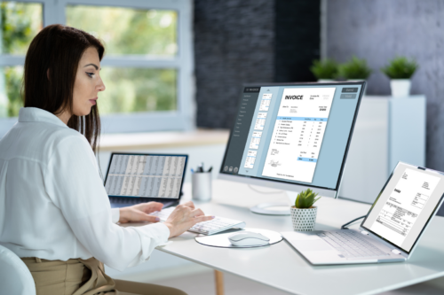 Femme assise à un bureau avec, devant elle, deux ordinateurs portables et un écran. Elle encode les factures électroniques de son entreprise dans un logiciel comptable sur son ordinateur.