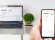 Mains tenant un smartphone avec, en arriière plan, un ordinateur portable ouvert sur un bureau, aux côtés d'une plante verte et d'une calculatrice. Sur les deux écrans est affiché une facture électronique notifiée comme étant payée, symbolisant la facturation électronique obligatoire dès janvier 2026 en Belgique.
