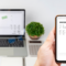 Mains tenant un smartphone avec, en arriière plan, un ordinateur portable ouvert sur un bureau, aux côtés d'une plante verte et d'une calculatrice. Sur les deux écrans est affiché une facture électronique notifiée comme étant payée, symbolisant la facturation électronique obligatoire dès janvier 2026 en Belgique.