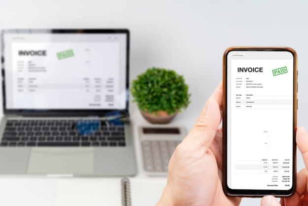 Mains tenant un smartphone avec, en arriière plan, un ordinateur portable ouvert sur un bureau, aux côtés d'une plante verte et d'une calculatrice. Sur les deux écrans est affiché une facture électronique notifiée comme étant payée, symbolisant la facturation électronique obligatoire dès janvier 2026 en Belgique.