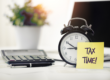 Une table de bureau avec, posés dessus, une plante verte, une calculatrice, un stylo-bille et un réveil sur lequel est collé un post-it indiquant "Tax Time!"