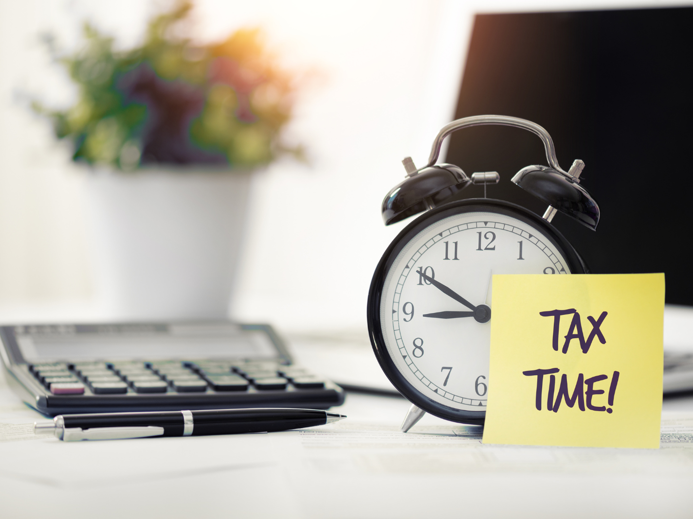 Une table de bureau avec, posés dessus, une plante verte, une calculatrice, un stylo-bille et un réveil sur lequel est collé un post-it indiquant "Tax Time!"